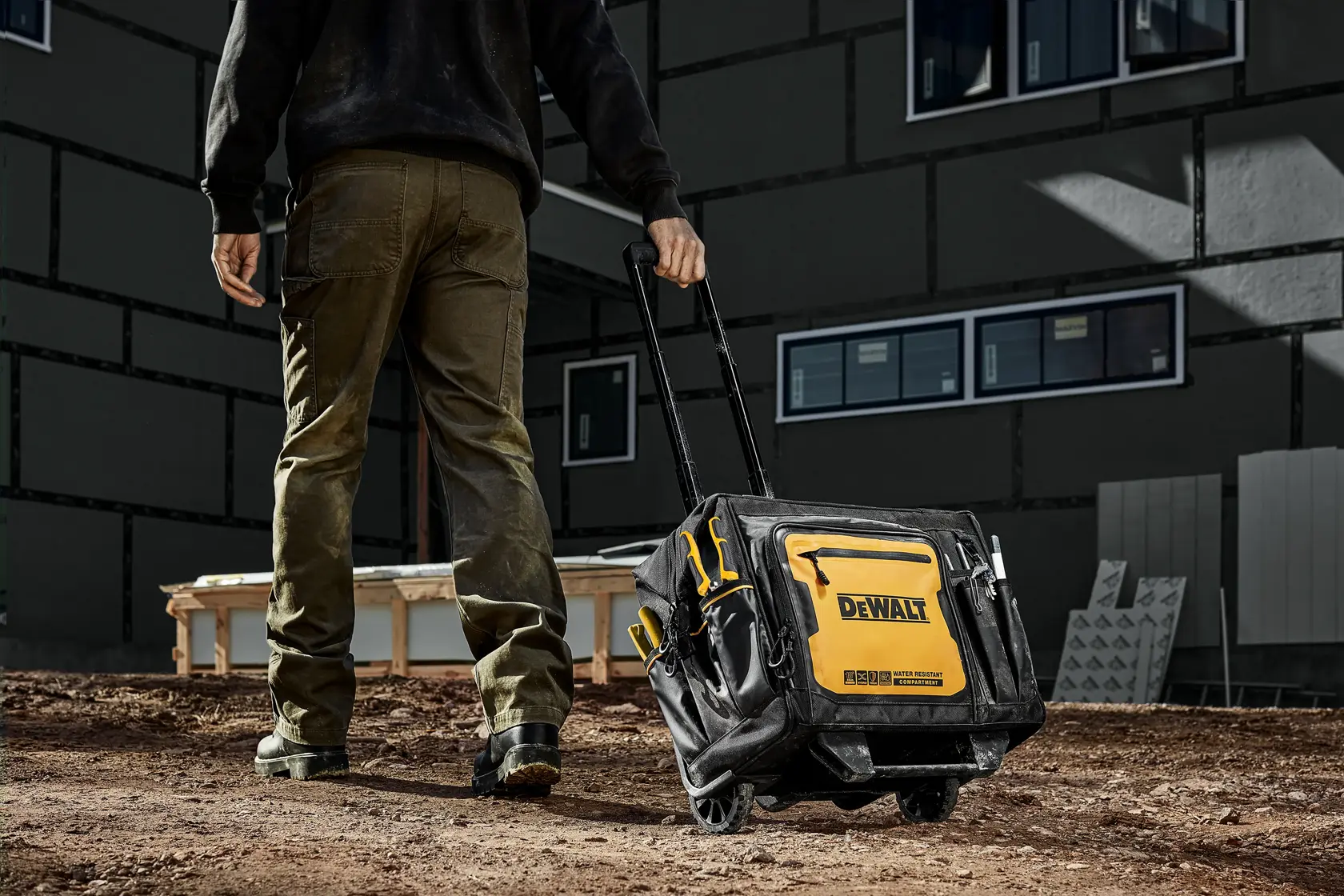 En person drar en DEWALT verktøybag med hjul og teleskophåndtak over en jordete byggeplass. Bagen er svart og gul, merket med DEWALT-logoen, og har flere rom for oppbevaring.