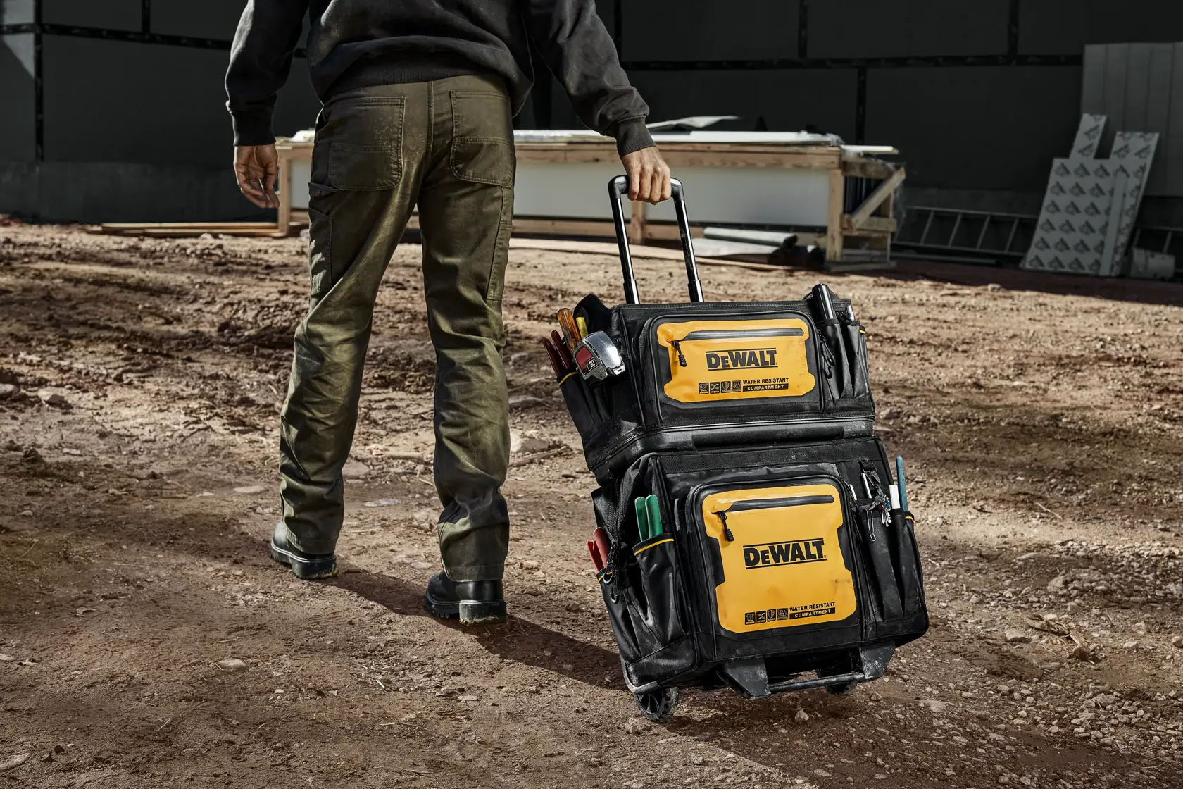 En person trekker en DEWALT verktøybag på hjul på en byggeplass. Baggen er svart og gul med flere lommer og rom, og er fylt med ulike verktøy.