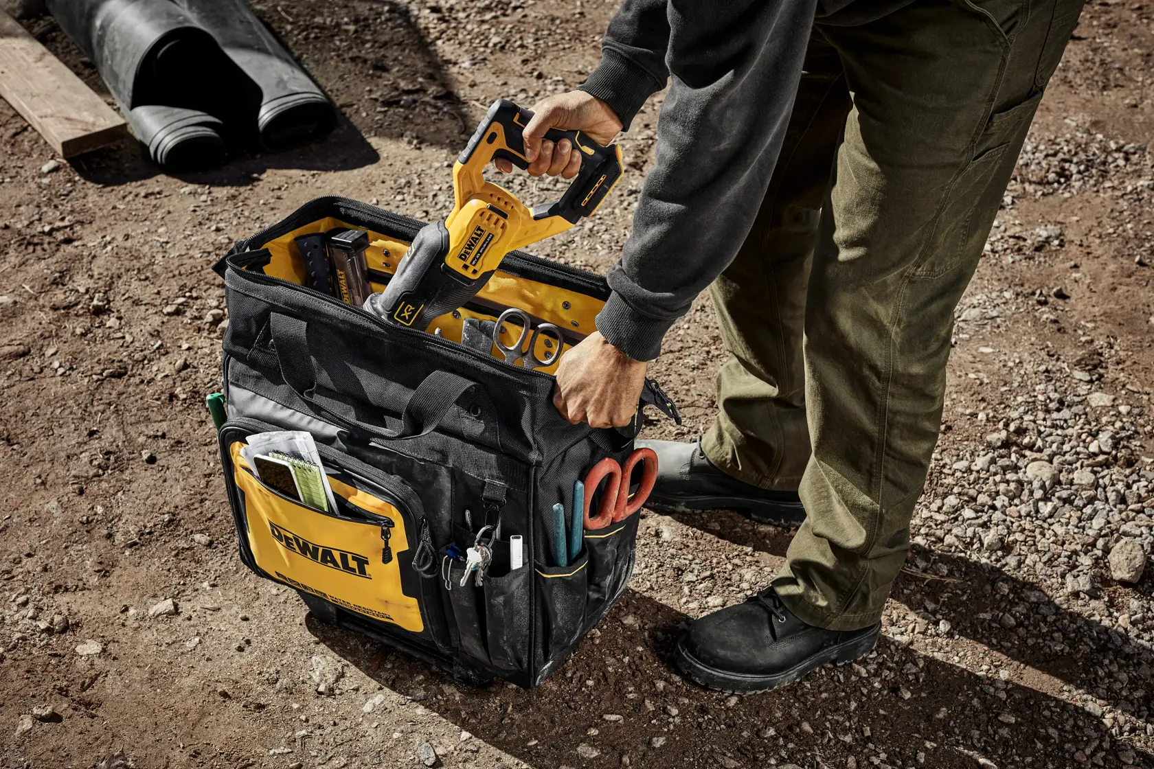En person plasserer et gult DEWALT elektrisk verktøy i en svart og gul DEWALT verktøyveske fylt med diverse verktøy og tilbehør, på jordgrunn på en utendørs arbeidsplass.
