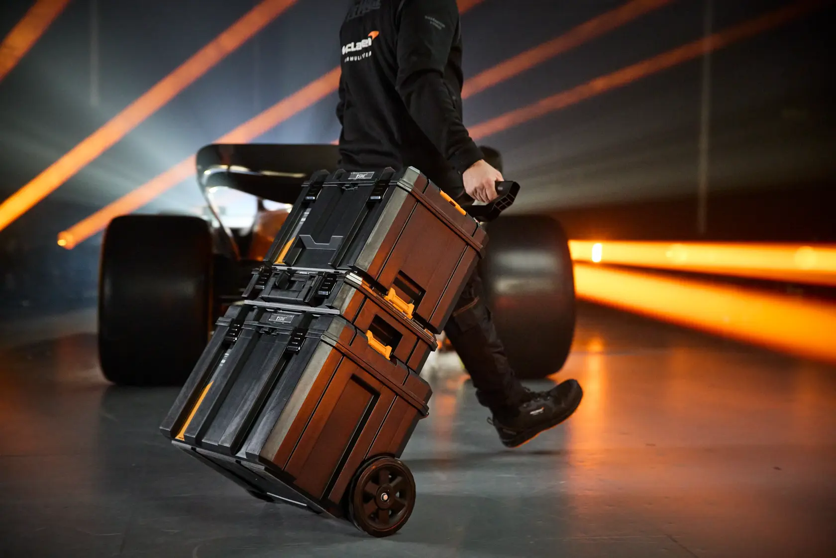 Person pulling stacked DEWALT wheeled toolboxes in a workshop with orange lights and a race car in the background.