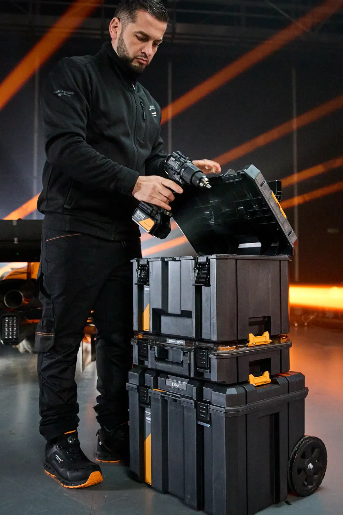 Person with blurred face holding a power tool near stacked DEWALT toolboxes with wheels in an indoor setting.
