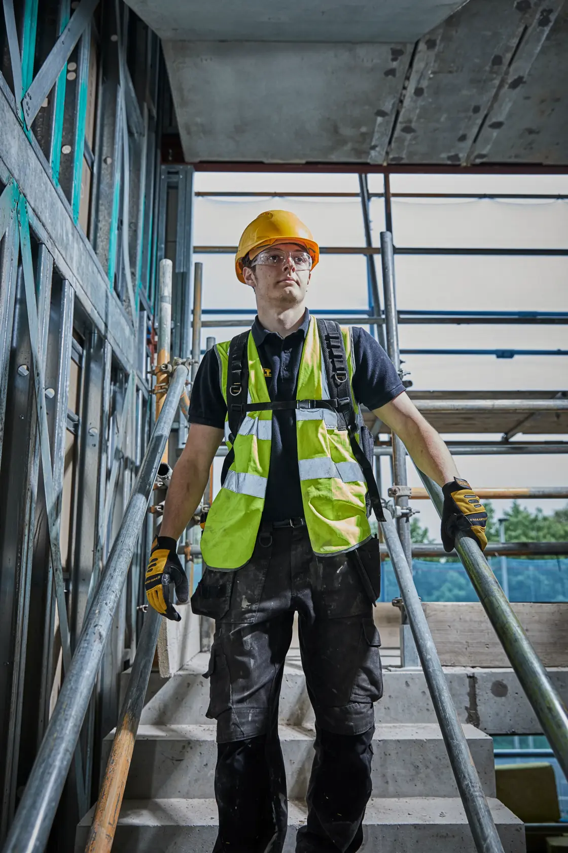 Persona con equipaggiamento di sicurezza e guanti su scale in cantiere con impalcatura metallica.
