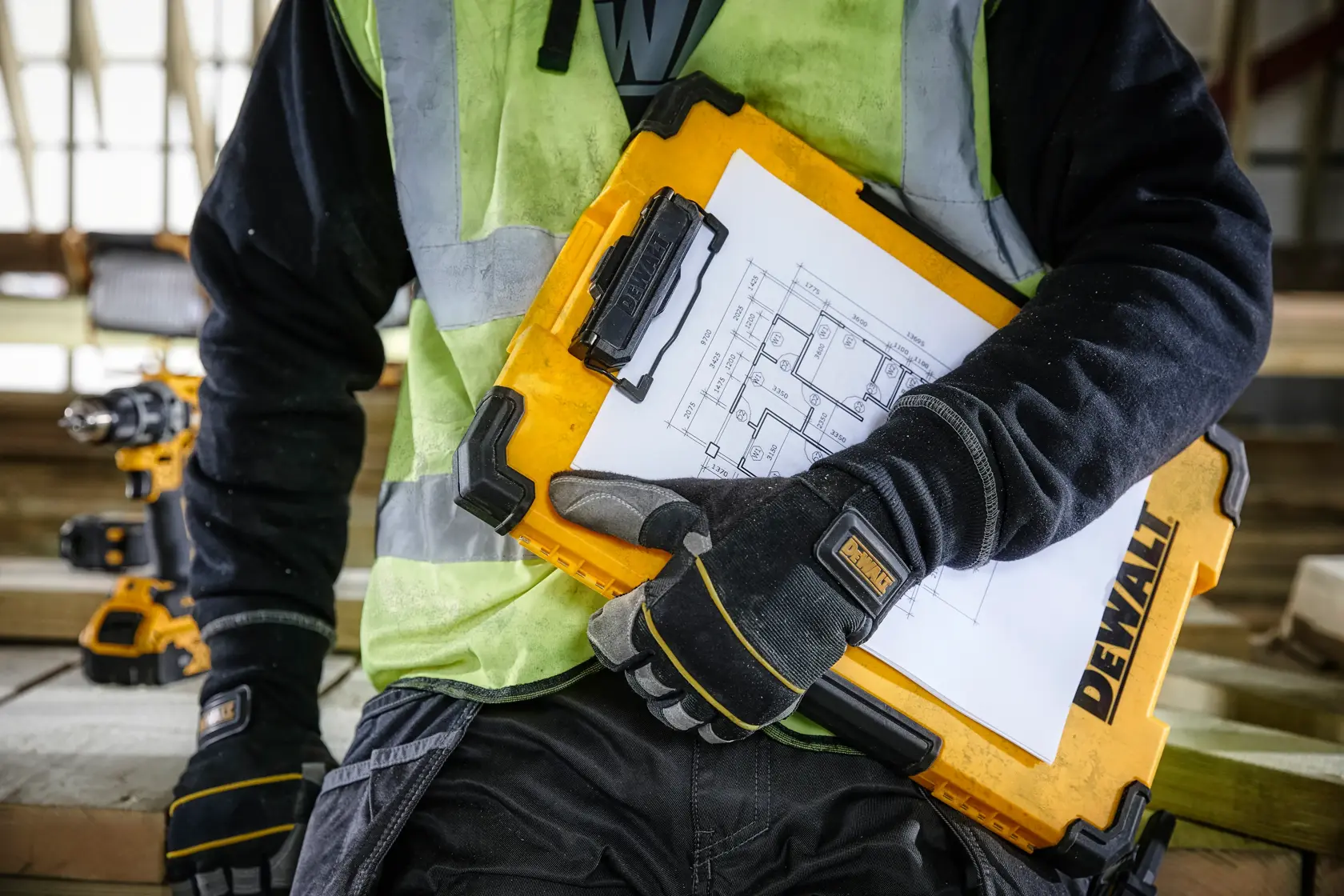 A person wearing work gloves and a high-visibility vest is holding a yellow DEWALT clipboard case with architectural drawings attached, at a construction site.