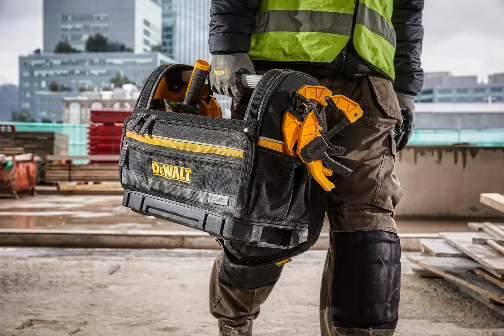 Person carrying a DEWALT tool bag with tools at a construction site, wearing protective gear and gloves.