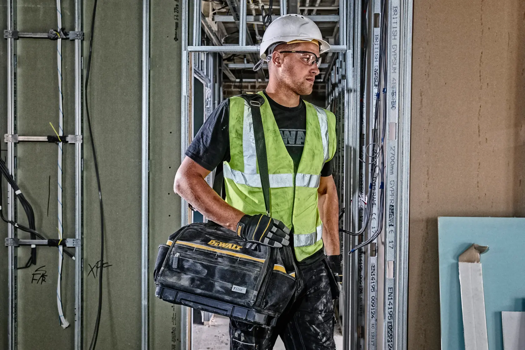 Worker in safety gear holding a DEWALT tool bag at a construction site with exposed walls and wiring.