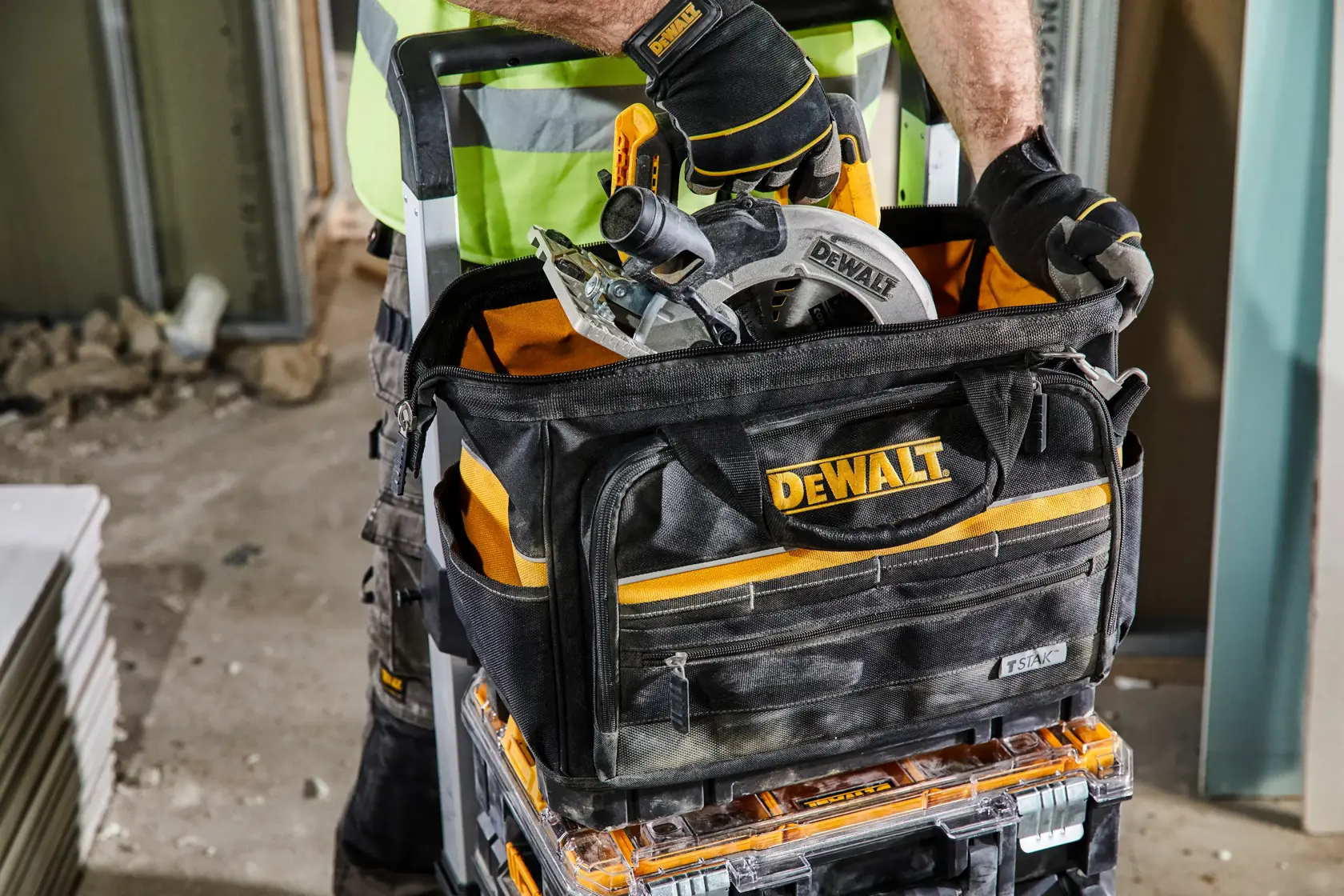 DEWALT tool bag with circular saw and tools on construction site. Person wearing gloves and safety vest handles equipment.