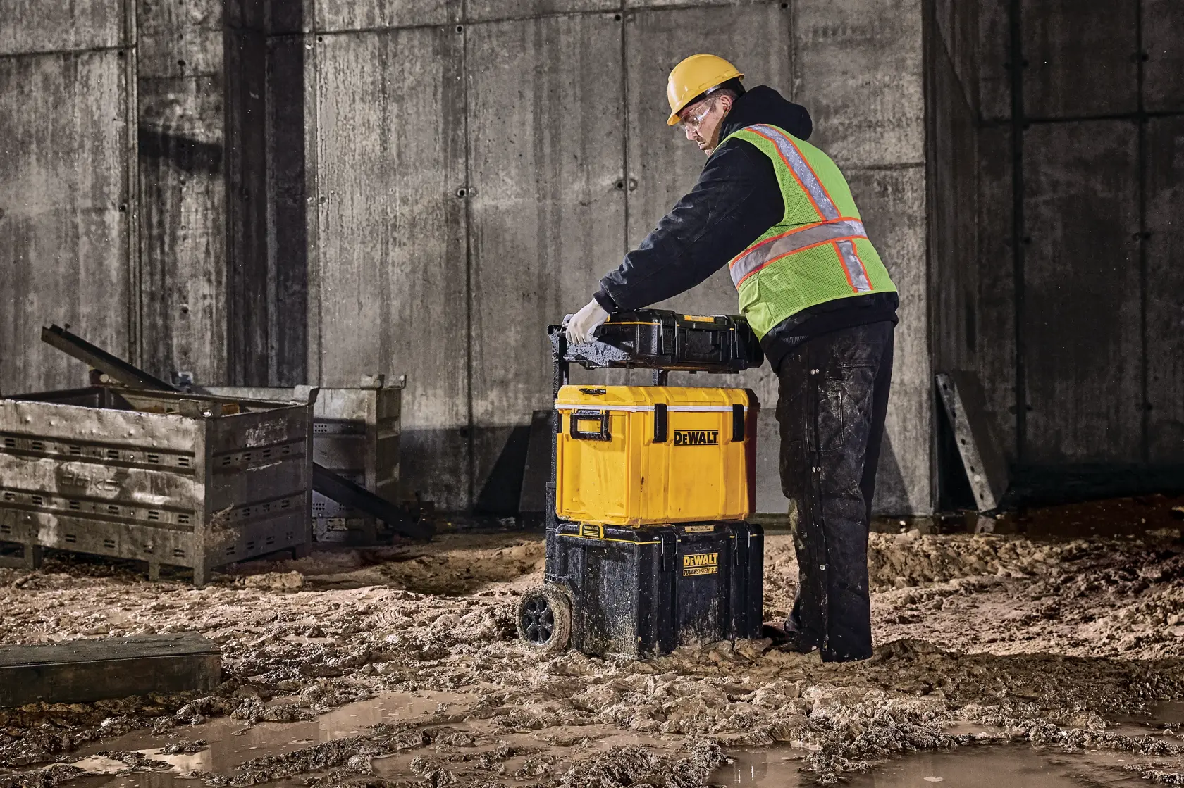 Person with safety gear uses DEWALT wheeled toolbox DWST83294-1 on a muddy construction site.