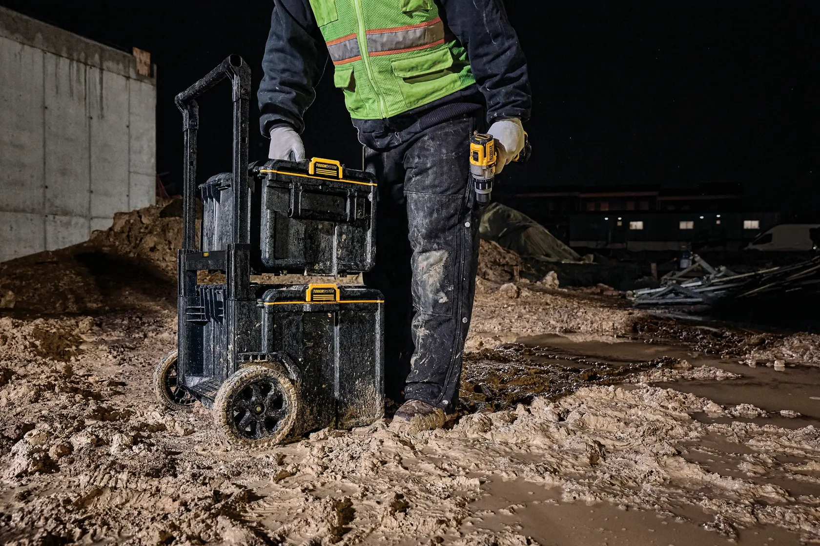 Person with DEWALT rolling tool box on muddy construction site at night, wearing safety gear and holding a drill.