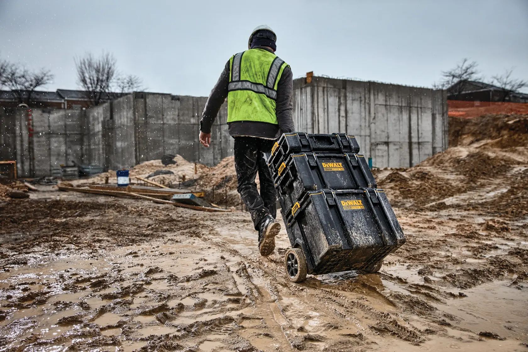 Worker pulls DEWALT toolboxes on wheels at muddy construction site, wearing safety gear and green vest.