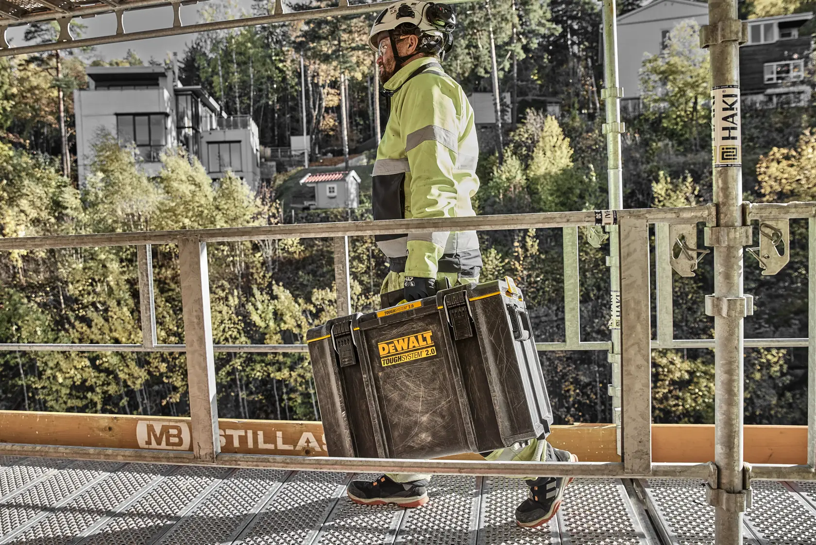 Person in safety gear carrying a DEWALT Toughsystem 2.0 toolbox on a construction site walkway.