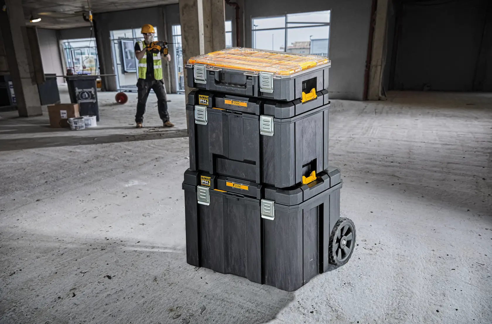 Stacked DEWALT toolboxes with wheels on a construction site floor, worker in background wearing safety gear.