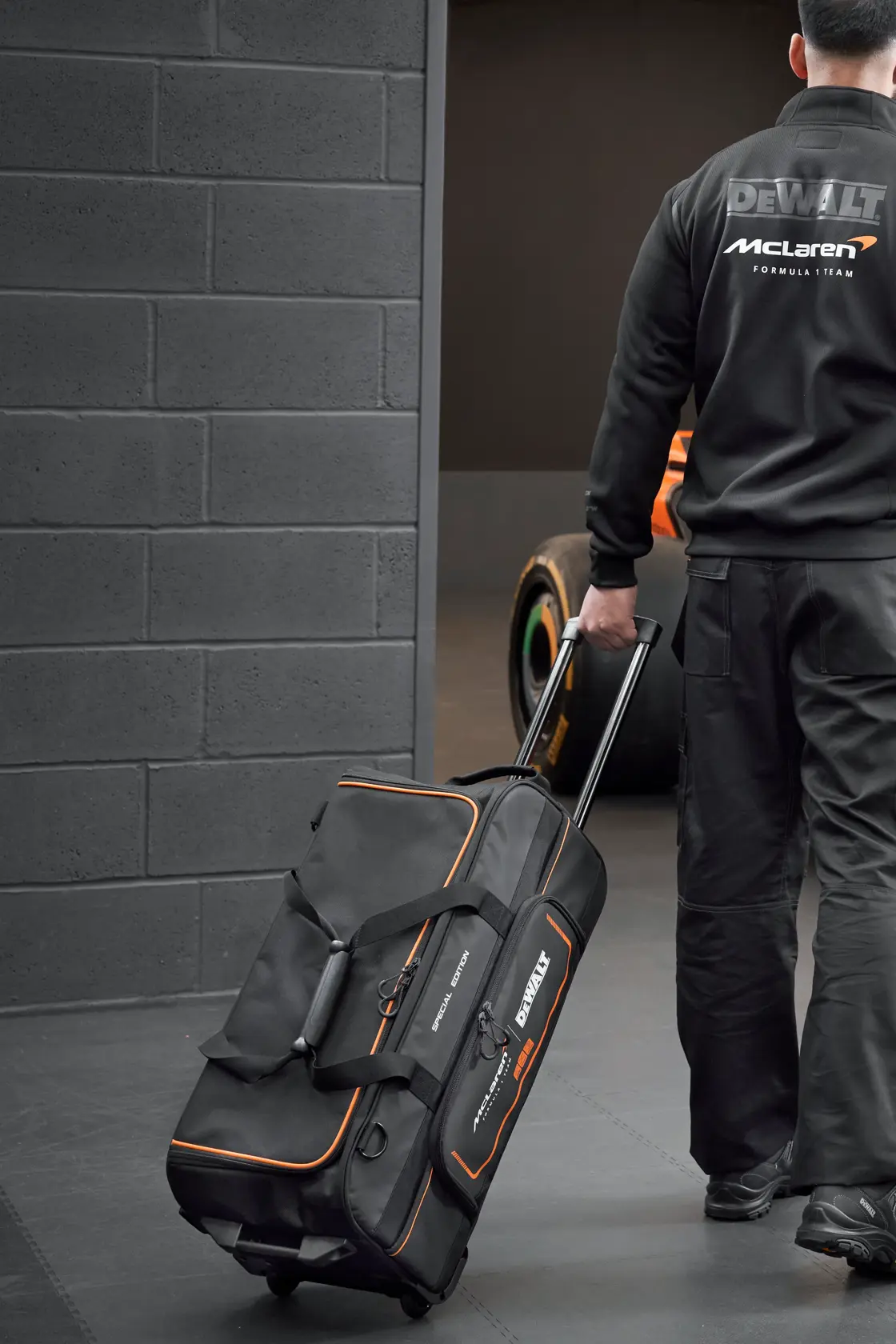 Person pulling a DEWALT McLaren wheeled tool bag in a garage with a race car visible in the background.