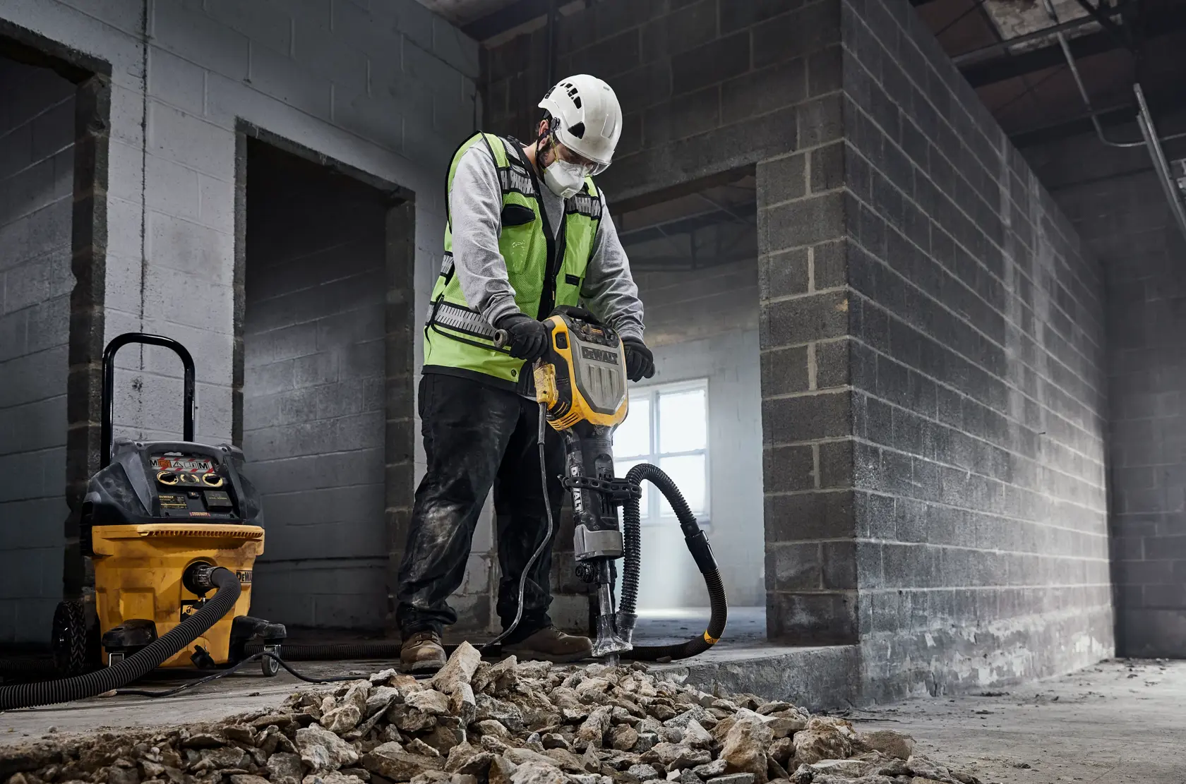 Worker uses DEWALT jackhammer and dust extractor on rubble inside a partially built industrial structure.