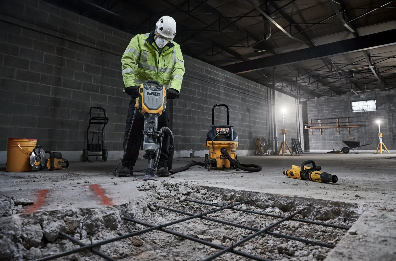 Worker uses DEWALT tool to break concrete in industrial setting, with dust extractor and other equipment nearby.